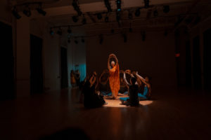 Five dancers dressed in a teal blue are sitting around a single, standing dancer in orange. All the dancers have their hands raised to the ceiling, forming a triangle between their fingers. Their surrounding is dark as the orange dancer is in a spotlight.