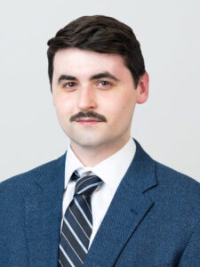 Owen Burnham, a fair-skinned man with brown-black hair and short mustache, sits in a blue textured suit, white shirt, and tie.