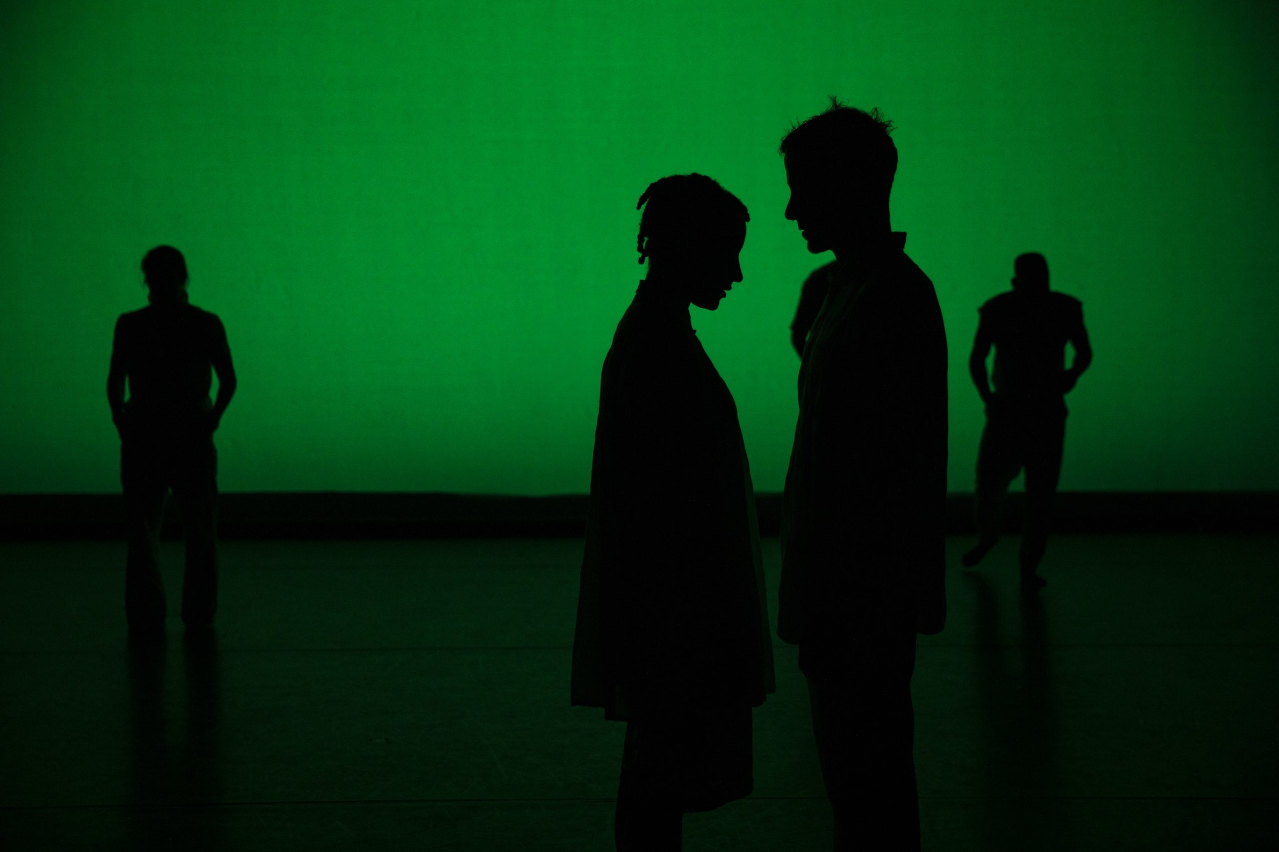 Four dancers - Kellie Ann Lynch, Myssi Robinson, Doug Gillespie, Jared Brown - silhouetted in green light stand in the frame. Two dancers, one furthest left and the other furthest right face the audience with heads tilted down. Two dancers in the center frame stand close together, facing each other in an inward moment.