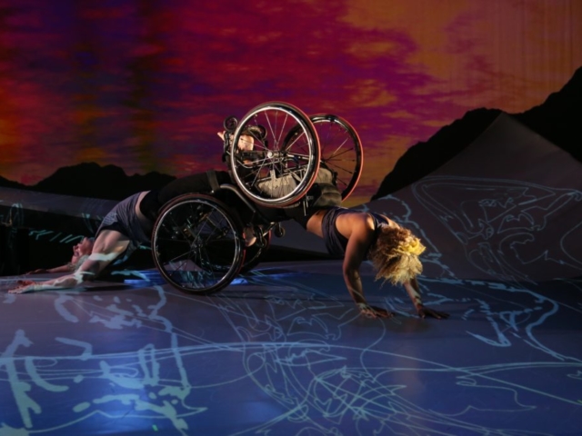 Alice Sheppard, a multiracial Black woman with coffee-colored skin and short curly hair, is crawling on her hands with her knees in Laurel Lawson's footplate. Laurel, a white dancer, is arching her back on the ground as she is dragged along the floor. A sunset appears behind them and shadowy figures appear below. Photo by MANCC / Chris Cameron.