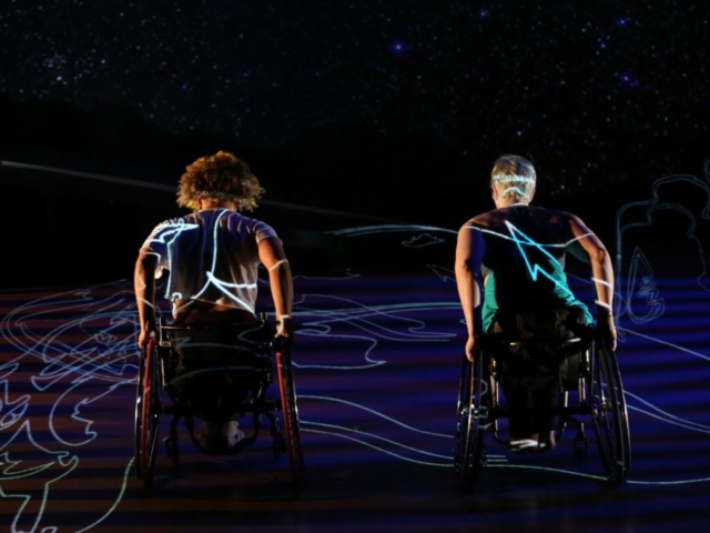 Alice Sheppard, a light-skinned multiracial Black woman, and Laurel Lawson, a pale white woman, push up the hill of the ramp, backs to the viewer and hands on their wheels, exerting effort. White lines of projected Rodin sculptures run from the deck of the ramp (which also features blue ripples of light) and across their backs. Photo by Chris Cameron/MANCC.⠀