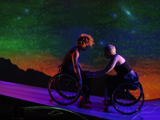 Against Michael Maag’s glowing green, gold, and blue starry sky, the ramp shines with lilac stripes and shadowy projections. Alice Sheppard, a light-skinned multiracial Black woman. wheels downhill, curly hair flying; she pushes Laurel Lawson, a white woman with very short cropped hair in a wheelchair, backwards. Laurel rests her hands on Alice's knees as they lock eyes.