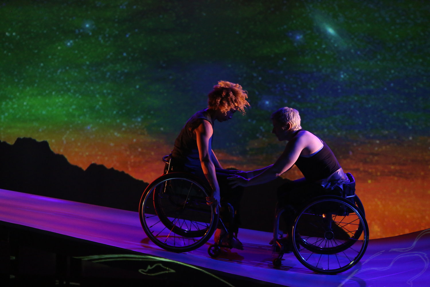 Against Michael Maag’s glowing green, gold, and blue starry sky, the ramp shines with lilac stripes and shadowy projections. Alice Sheppard, a light-skinned multiracial Black woman. wheels downhill, curly hair flying; she pushes Laurel Lawson, a white woman with very short cropped hair in a wheelchair, backwards. Laurel rests her hands on Alice's knees as they lock eyes.