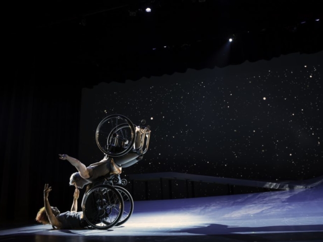 Laurel Lawson, a white dancer, balances on Alice Sheppard’s footplate, with arms spread wide, wheels spinning. Alice, a multiracial Black woman with coffee-colored skin, opens her arms wide to receive her in an embrace. A starry sky fills the background, and moonlight glints off their rims. Photo by BRITT / Jay Newman.