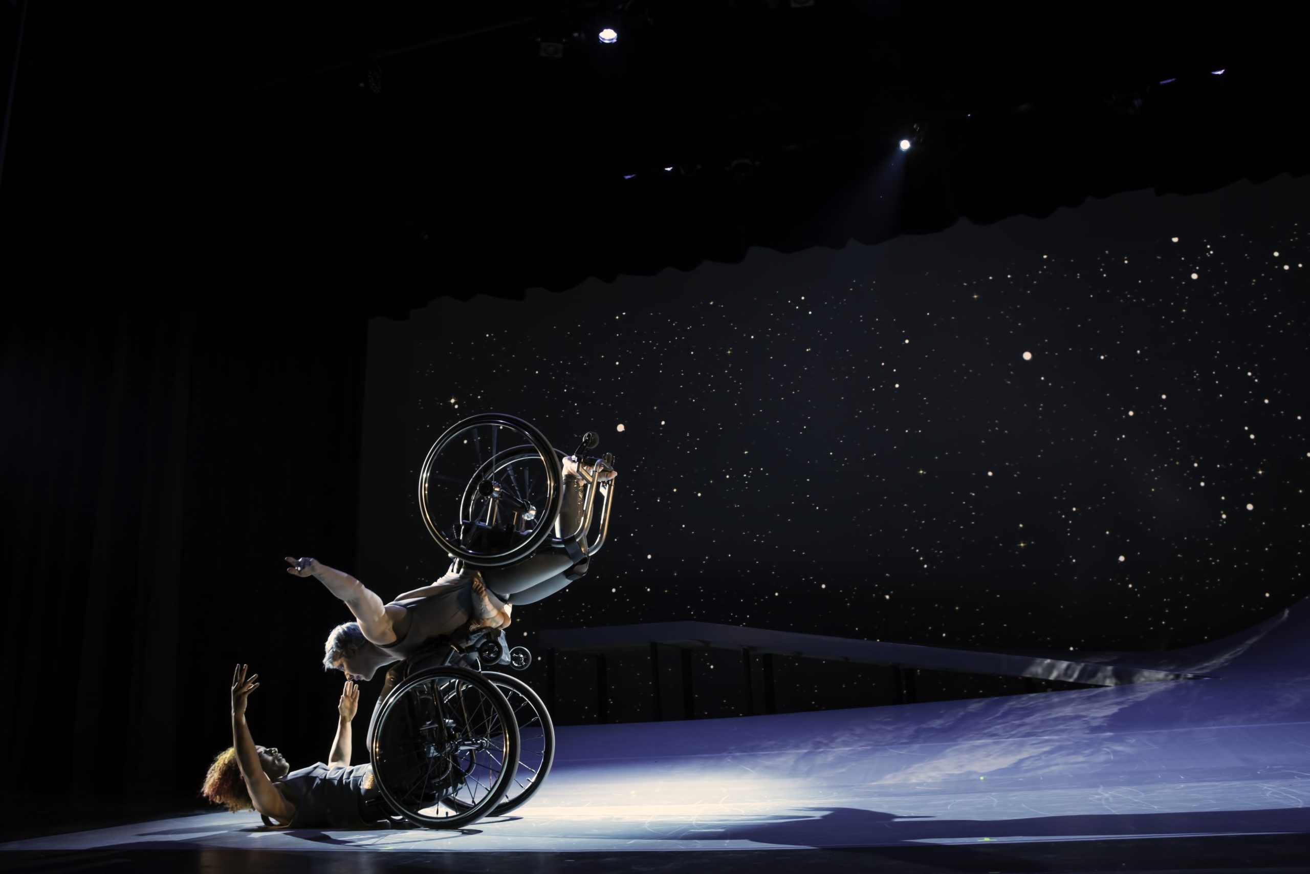 Laurel Lawson, a white dancer, balances on Alice Sheppard’s footplate, with arms spread wide, wheels spinning. Alice, a multiracial Black woman with coffee-colored skin, opens her arms wide to receive her in an embrace. A starry sky fills the background, and moonlight glints off their rims. Photo by BRITT / Jay Newman.