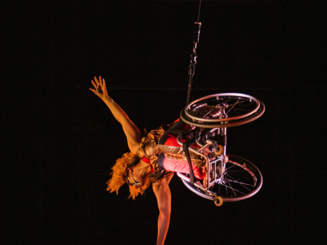 Alice Sheppard flies in her wheelchair, body parallel to the stage as she reaches her arms wide, one hand almost skimming the floor. She is a multiracial Black woman with coffee colored skin and short curly hair; she wears a red and gold bodysuit, black and white lines adorn her face.