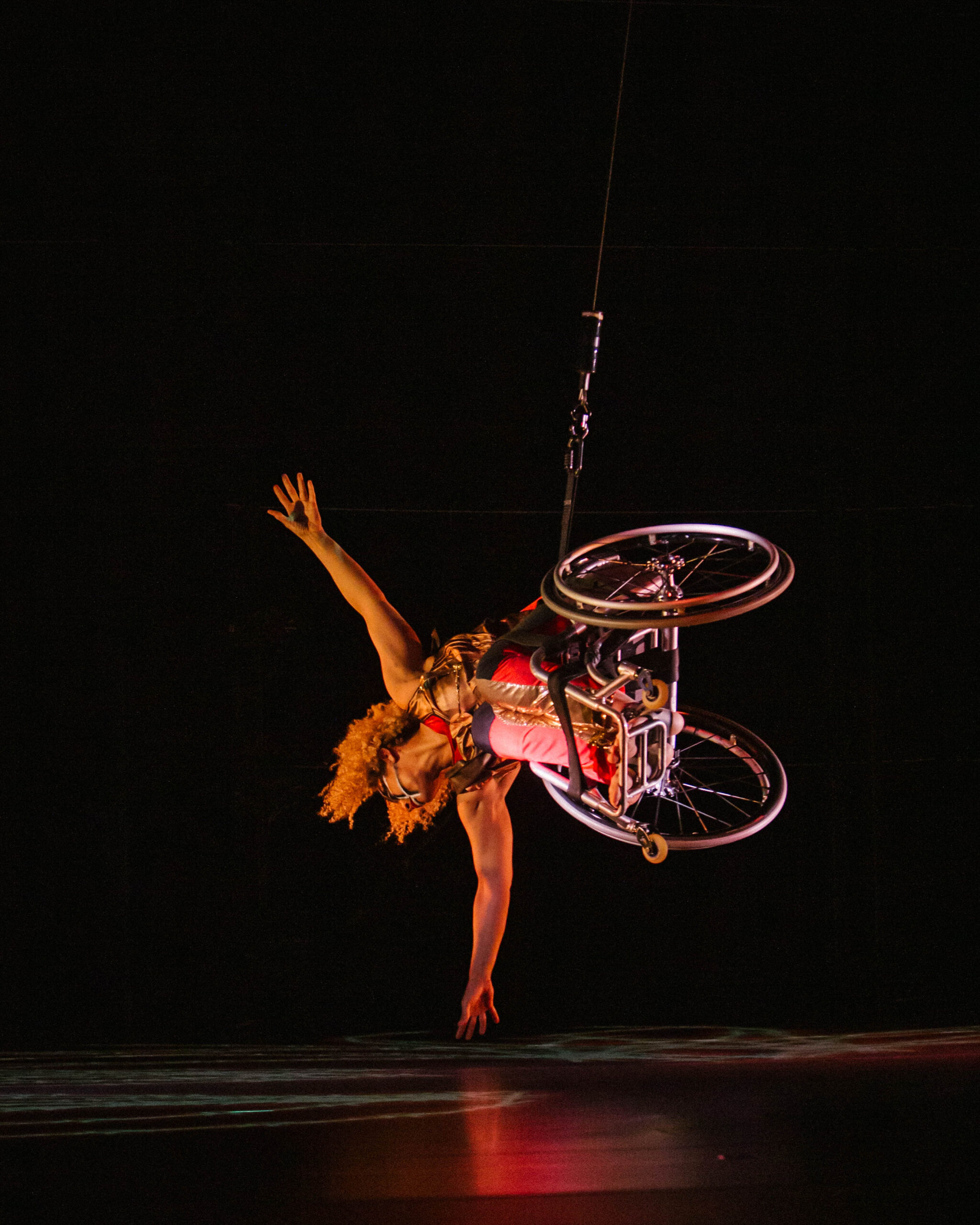 Alice Sheppard flies in her wheelchair, body parallel to the stage as she reaches her arms wide, one hand almost skimming the floor. She is a multiracial Black woman with coffee colored skin and short curly hair; she wears a red and gold bodysuit, black and white lines adorn her face.