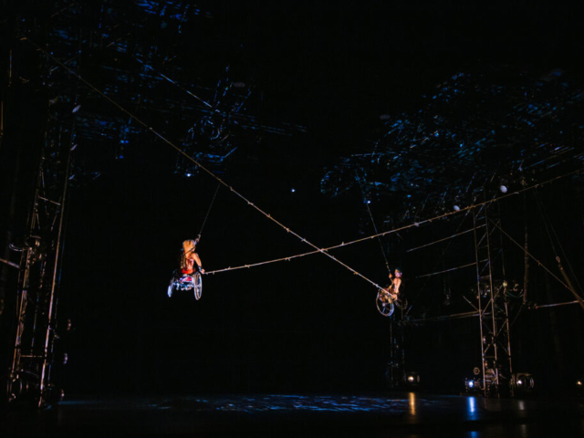 Alice Sheppard and Laurel Lawson fly through the air. They look small amidst the large, dark performance space. Barbed wire extends and crosses between them, creating a big X, as their chairs and the tall metal truss that surrounds them shine in the theatrical light. The expansive ceiling above them is spackled in blue projection.