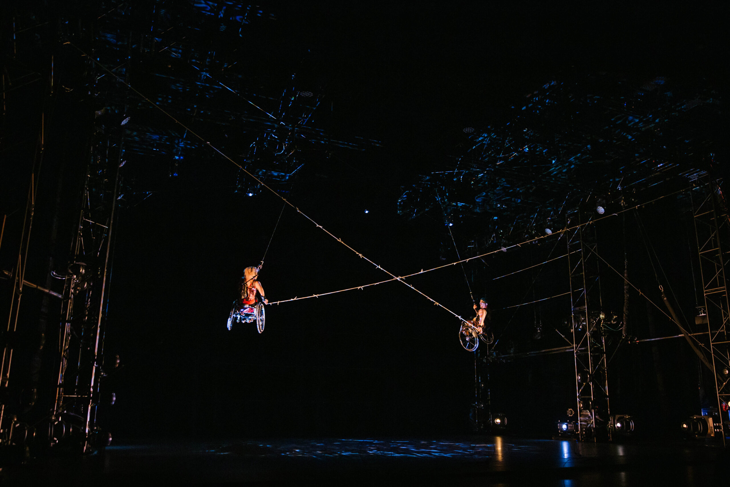 Alice Sheppard and Laurel Lawson fly through the air. They look small amidst the large, dark performance space. Barbed wire extends and crosses between them, creating a big X, as their chairs and the tall metal truss that surrounds them shine in the theatrical light. The expansive ceiling above them is spackled in blue projection.