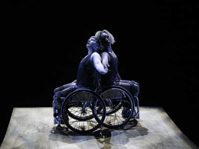 Cast in a shadowy blue overhead light, Laurel Lawson and Alice Sheppard are back to back on a wooden panel with a black background. Alice is a light-skinned multiracial Black woman with short curly hair and Laurel Lawson is a white woman with short cropped hair. Both dancers extend their arms toward the camera, with Alice gazing towards her fingers and Laurel gazing upward, eyes closed.