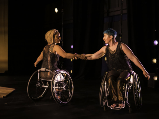 With stage lights in the background, dancers Alice Sheppard and Laurel Lawson look at each other with their arms interlaced, their bodies an arm length apart, yet facing opposite directions. Laurel, a white woman with cropped blue hair, faces toward the camera, with Alice: a light-skinned multiracial Black woman, faces away. They are dressed in shimmering autumnal tones.