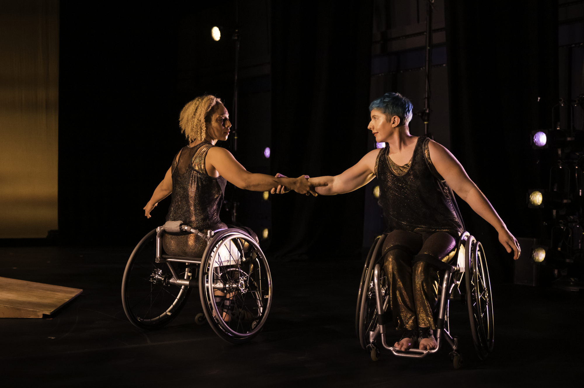 With stage lights in the background, dancers Alice Sheppard and Laurel Lawson look at each other with their arms interlaced, their bodies an arm length apart, yet facing opposite directions. Laurel, a white woman with cropped blue hair, faces toward the camera, with Alice: a light-skinned multiracial Black woman, faces away. They are dressed in shimmering autumnal tones.