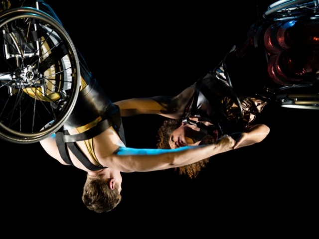 A close up of Alice Sheppard and Laurel Lawson suspended in the air, arms outstretched and clasping each other’s hands. Alice is a multiracial Black woman with coffee-colored skin and short curly hair; she wears a shimmery deep red costume. Laurel is a white dancer with very short cropped hair; she wears a shimmery gold costume with thick black shoulder straps. The dancers are somehow upside down and horizontal at the same time, their wheels shining and facing out; if they let go, they will swing like pendulums. Photo Robbie Sweeny/Kinetic Light.