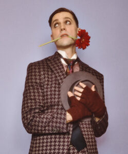 A man, wearing a burgundy and grey houndstooth suite, glances upward with a red flower in his teeth. He holds a grey brimmed hat against his heart with burgundy fingerless gloves. The background is lavender.