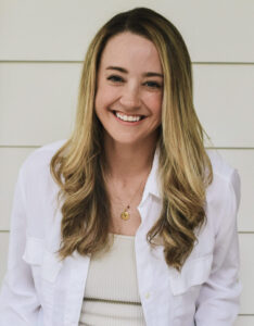 Kelsey, a female with long blonde/brown hair, sitting and smiling with a white button down shirt on.