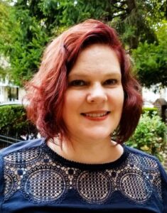 A white woman with red hair smiles at the camera. In the background is vibrant green foliage and she wears a navy blue shirt with circular designs.