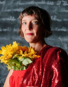 Photo by Karla Conrad. White Woman with brown hair in red dress with smudged out blackboard behind her. She is holding a big bunch of yellow sunflowers to her chest and it comes out the front of her dress. This photo is from Esther's performance with Propelled Animals at Grinnell College, Iowa.