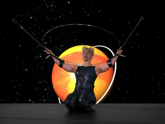 Alice Sheppard kneels, holding a huge silver ring and crutches high. Leaning back, she casts her gaze up toward the ring’s glinting edge. Behind her, a video projection of the planet Saturn, burning with bright yellows and oranges and featuring its own celestial ring, surrounded by stars. Alice is a multiracial Black woman with short curly blonde hair piled atop her head; she wears a dark shimmery sleeveless bodysuit.