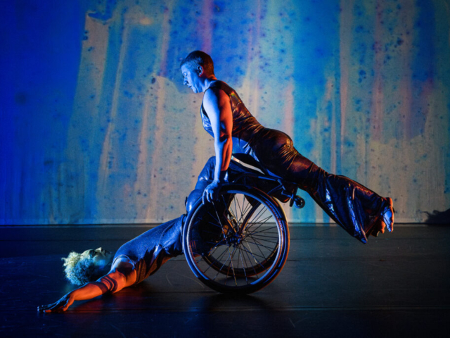Laurel Lawson holds their body in a diagonal line as they balance atop Alice Sheppard’s wheels and shins. Alice supports from below as they lay back, arms and torso on the ground, hips lifted. Alice is a multiracial Black woman with short curly hair, Laurel is a white person with very short cropped hair; they both wear shimmery sleeveless bodysuits. Behind them, a projection of soft blues, purples, and greens looks like rain on a windowpane.