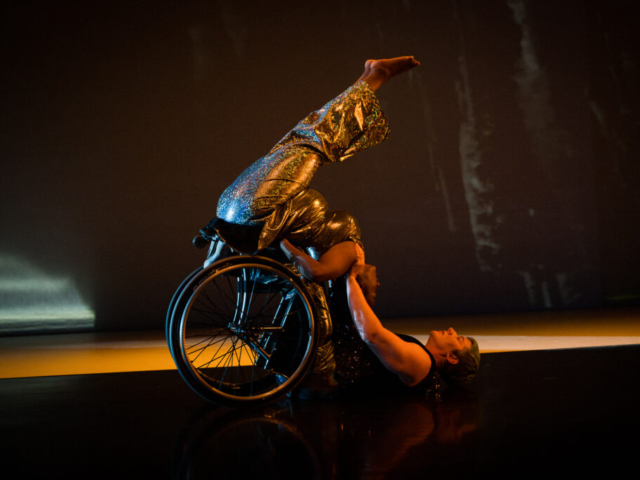 Laurel Lawson partners Kayla Hamilton as they lay with their back on the floor, hands holding Kayla’s shoulders, and legs and wheels supporting Kayla’s lower body. Kayla balances as she tucks her torso and lifts her hips and extended legs, making a diagonal line with her lower body. Laurel is a white person with very short hair wearing a black sleeveless top. Kayla is a thick bodied Black woman wearing a silver sleeveless top and matching pants.