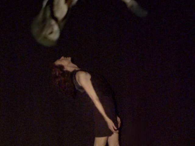 Myrna Packer stands alone at center stage, leaning back with her chest open and head tilted upward. Above her, an image of her younger self—taken from decades-old archival footage—appears suspended in midair, flipped upside down in a dramatic arc.