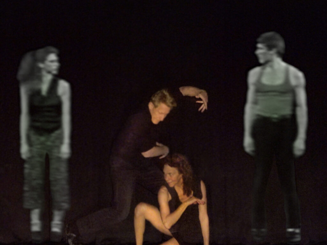 Art Bridgman and Myrna Packer are in the center of the photo. Bridgman is standing with his body curved over Packer who is crouched on the floor looking toward the right. Images of them from archival video footage from decades ago frame them, creating a composition that melds present and past.